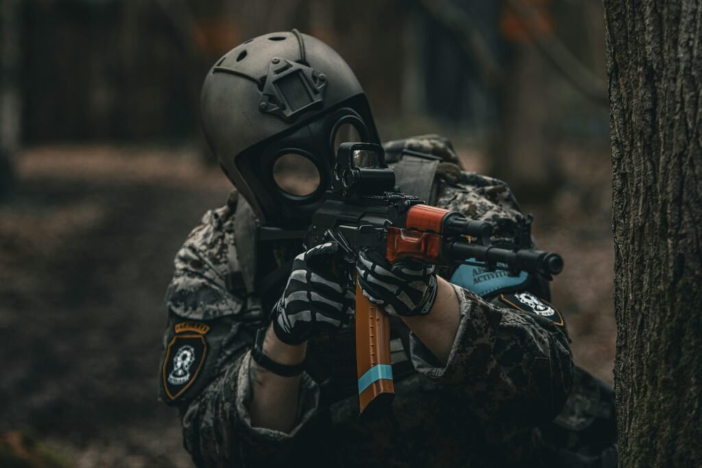 Paintball player in full gear and gas mask aiming a paintball gun outdoors.